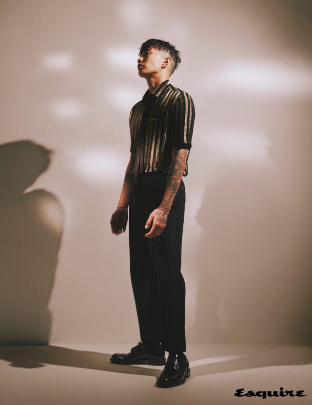 골드 스트라이프 쇼트 슬리브 폴로셔츠, 트리플 플리츠 하이웨이스트 팬츠, 아미 부츠, 타이, 팔찌 모두 생 로랑 by 안토니 바카렐로.