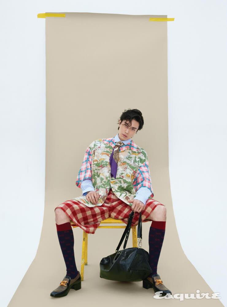 멀티컬러 펠라인 프린트의 오버사이즈 볼링 셔츠, 하늘색 스트라이프 셔츠, 울 매크로 오버사이즈 쇼츠, GG 패턴 양말, 체인 호스빗 디테일의 리저드 가죽 소재 부티, 인터로킹 G 디테일과 사이드 핸들이 특징인 스몰 더플백, 실크 스카프 모두 구찌.